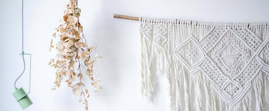 A white wall decorated with a mint green pendant light, a hanging bundle of dried eucalyptus leaves, and a large white macramé tapestry with geometric diamond patterns.