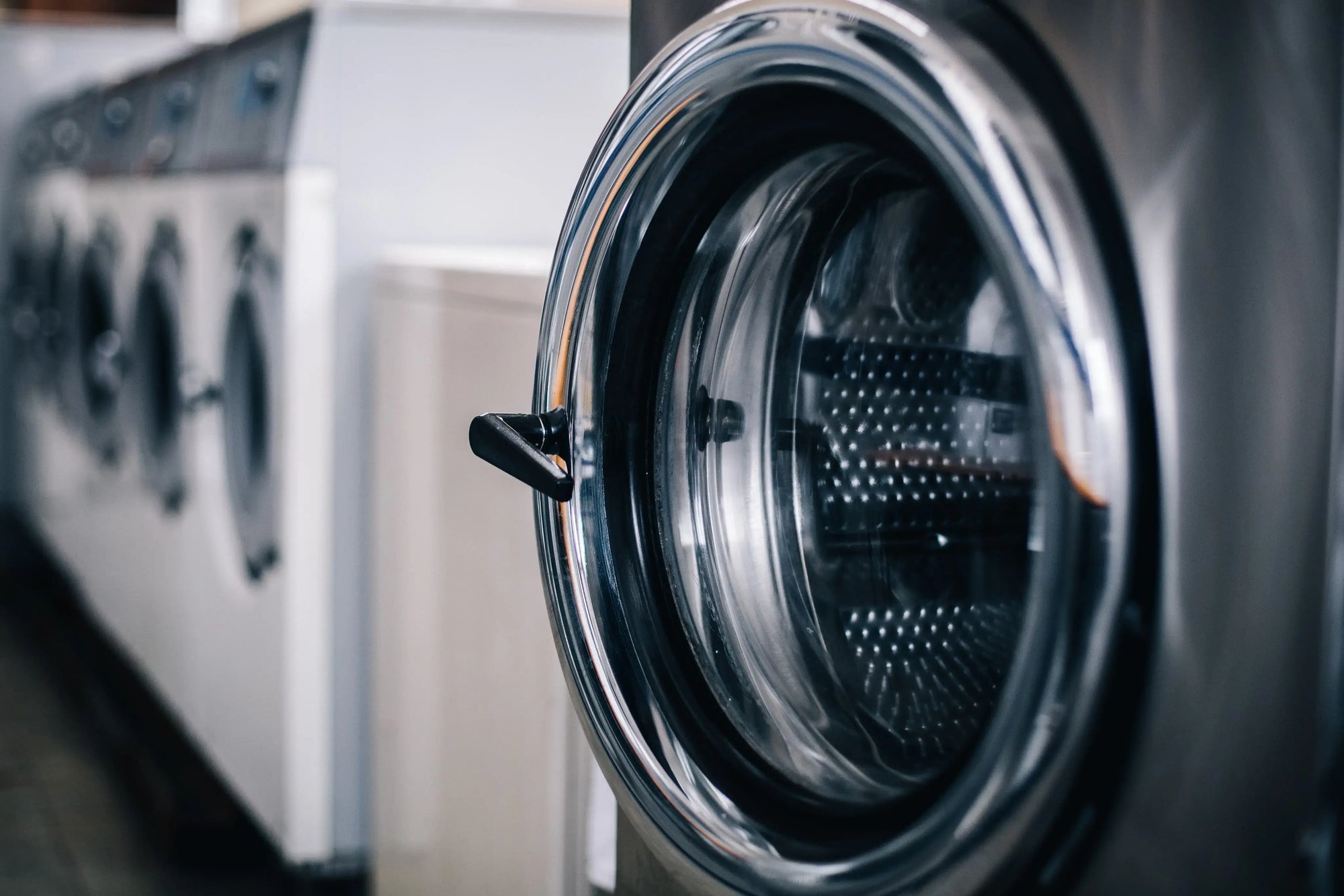 Stainless steel front-load washing machine with other laundry appliances blurred in a laundry room.