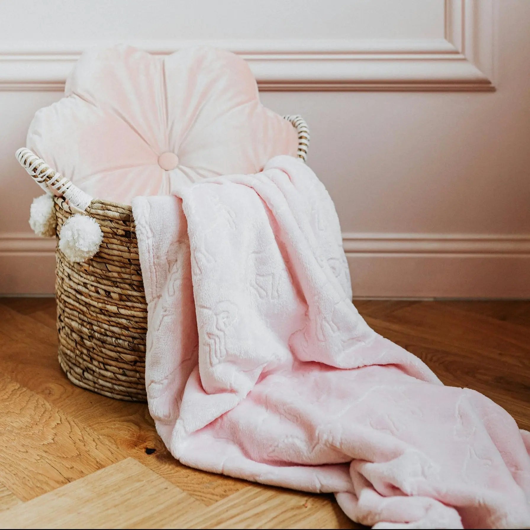 A woven wicker cat bed basket decorated with white pom-poms, sitting on a wooden herringbone floor. It contains a tufted pink velvet cushion and a soft pink fleece blanket draped over the side.